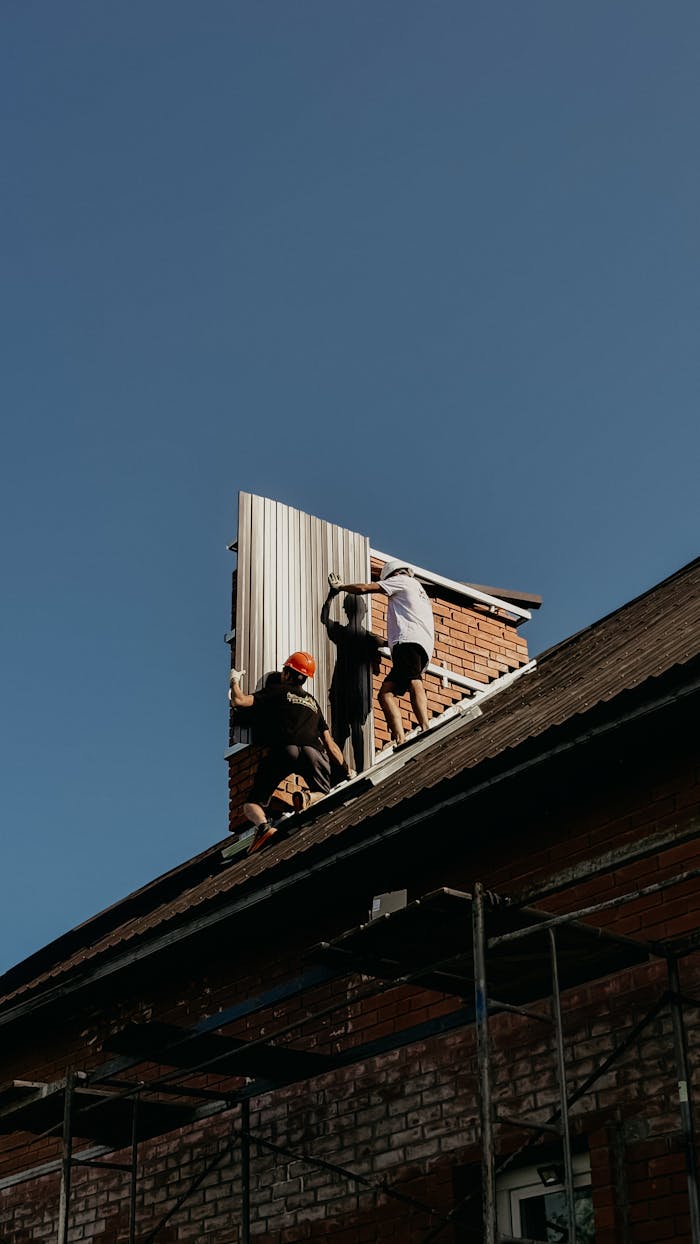 Three workers installing metal roof panels on a brick house under clear skies.